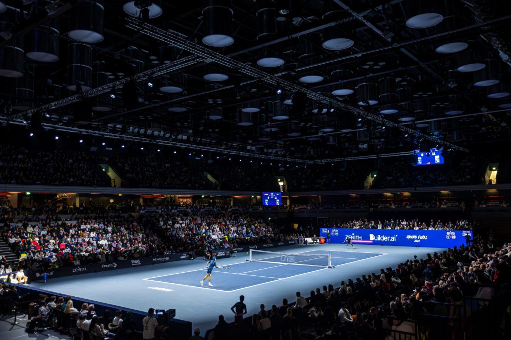 Professional tennis match taking place in a large indoor arena with spectators, LED screens and event lighting.